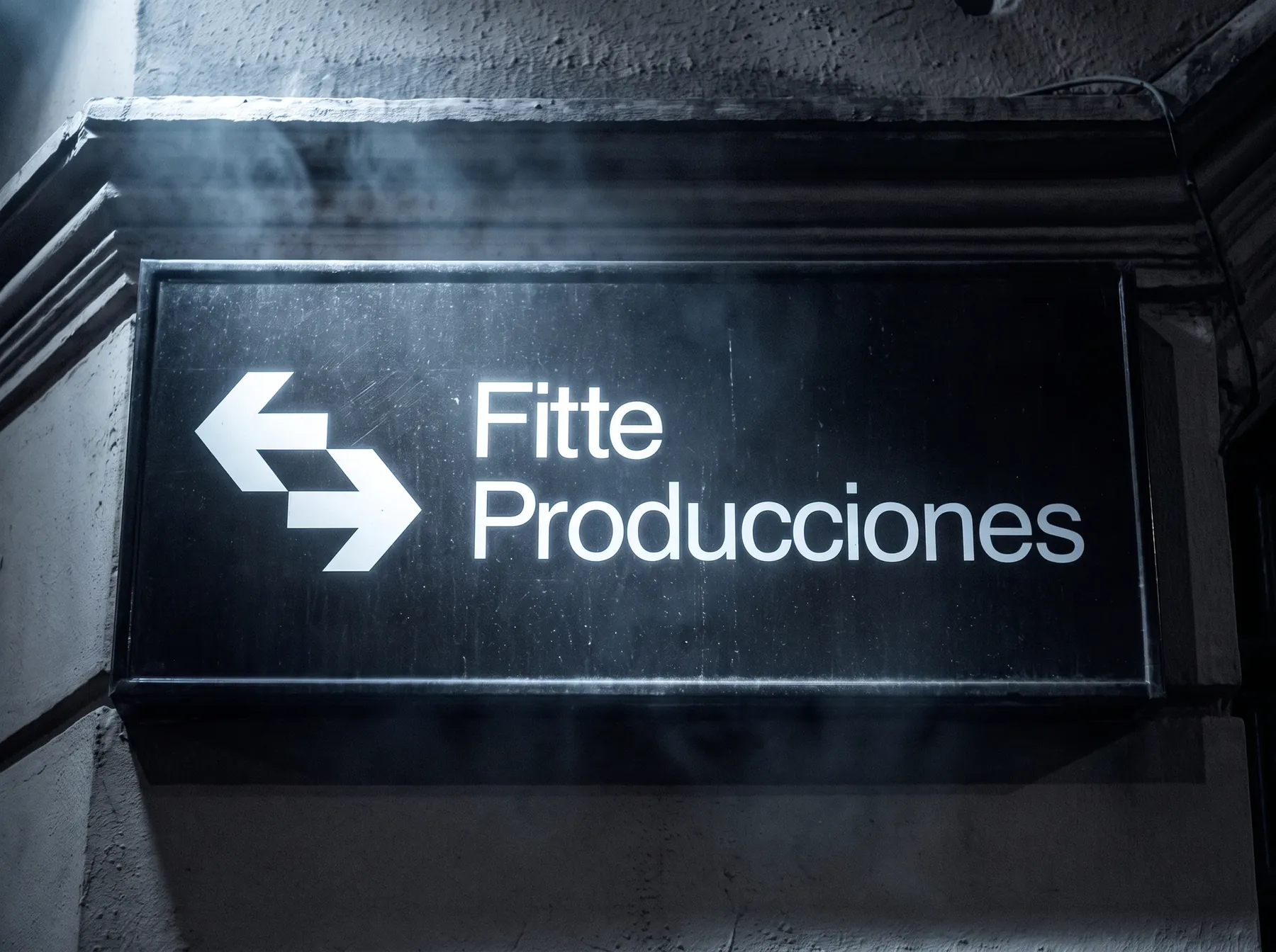Cartel luminoso con el logo de flechas y el nombre Fitte Producciones, colgado en una pared de noche.