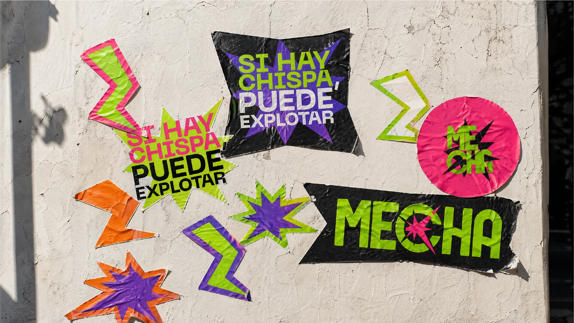 Stickers de Mecha con el logo, rayos y el lema "Si hay chispa, puede explotar", pegados en una pared de la calle.